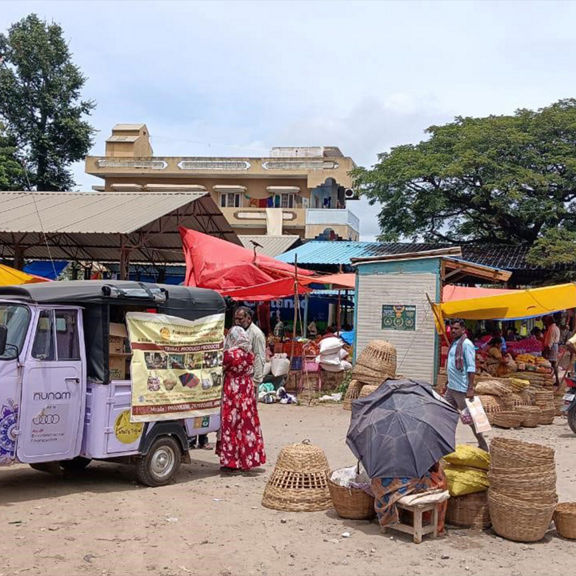A lilac electric rickshaw is parked at a busy market with vendor stalls and pedestrians.