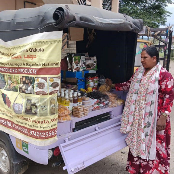A woman sells groceries from a lilac electric rickshaw with a promotional banner and open sales compartment.