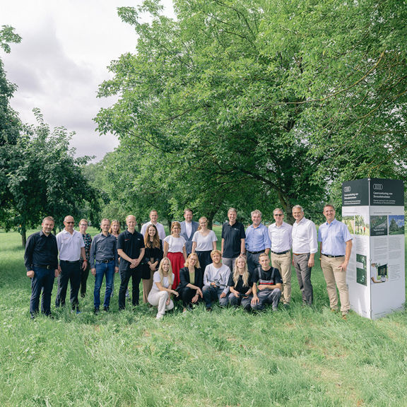 A group of 20 people stand in an orchard next to an information board and look at the camera together.