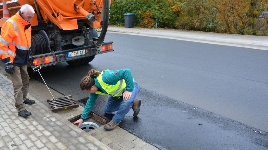 Zwei Männer an einem offenen Gullydeckel, einer kniet und prüft die Öffnung, ein orangefarbener LKW steht daneben.