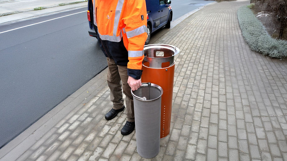 Mann in Warnjacke steht auf Gehweg und hält ein zylindrisches Metallsieb neben einem offenen Filtersystem.