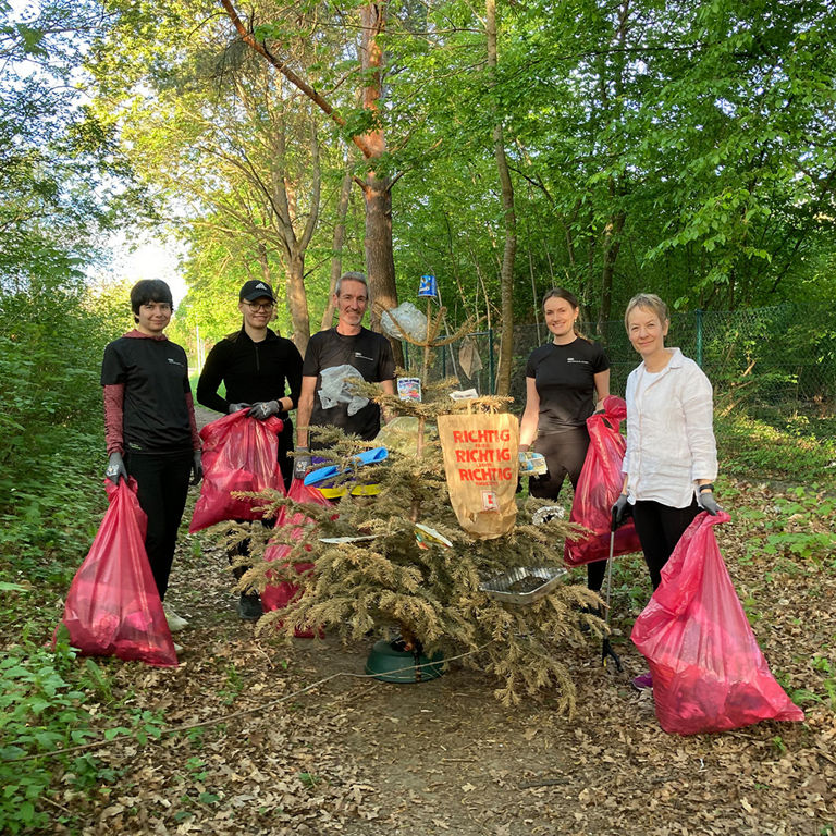 Six people in a wooded area holding pink trash bags, standing beside a pile of collected litter.