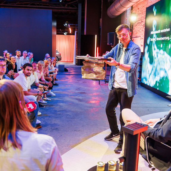 Moderator zeigt dem Publikum ein rundes Filterelement, viele Personen sitzen gegenüber und hören zu.