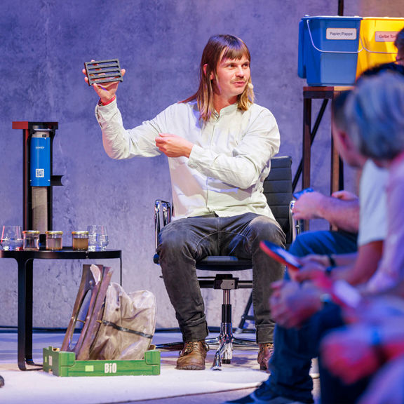 Ein Mann mit langen Haaren zeigt dem Publikum ein rechteckiges Objekt, im Hintergrund Recycling-Boxen.