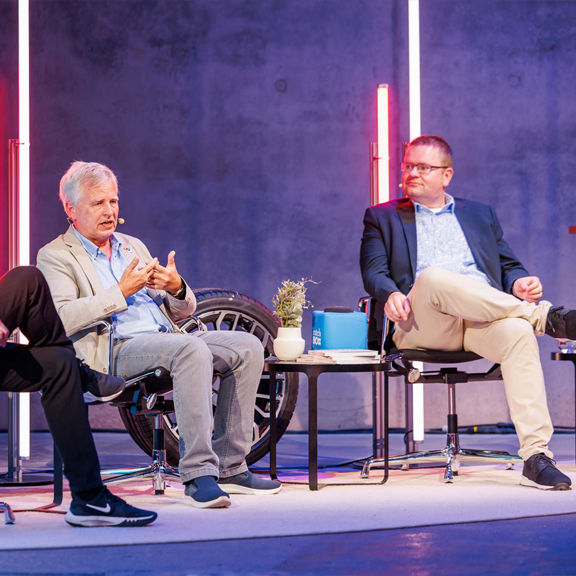 Drei Männer diskutieren auf einer Bühne über Mikroplastik, im Hintergrund leuchten farbige Lichtstäbe.