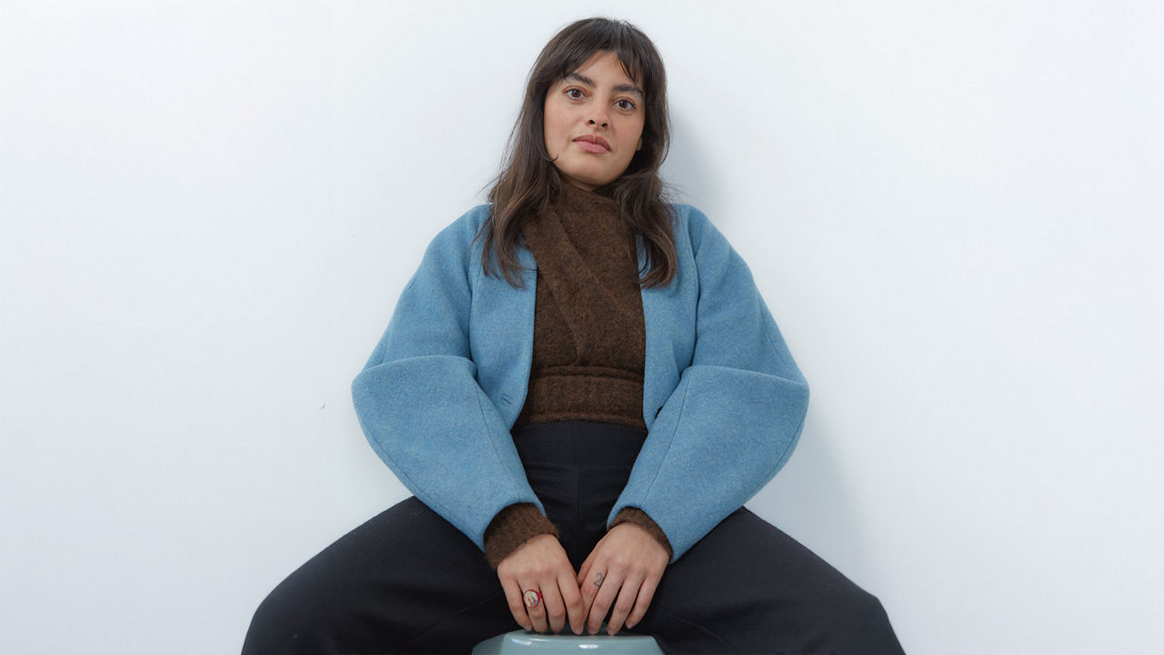 Frau mit mittellangen dunklen Haaren im braunen Pullover und blauer Jacke, vor weißem Hintergrund sitzend.