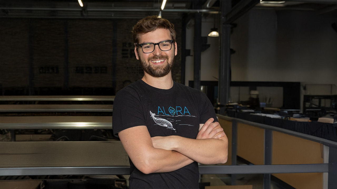 Mann mit Bart und Brille im schwarzen T-Shirt, Arme verschränkt, in einer Industriehalle stehend.