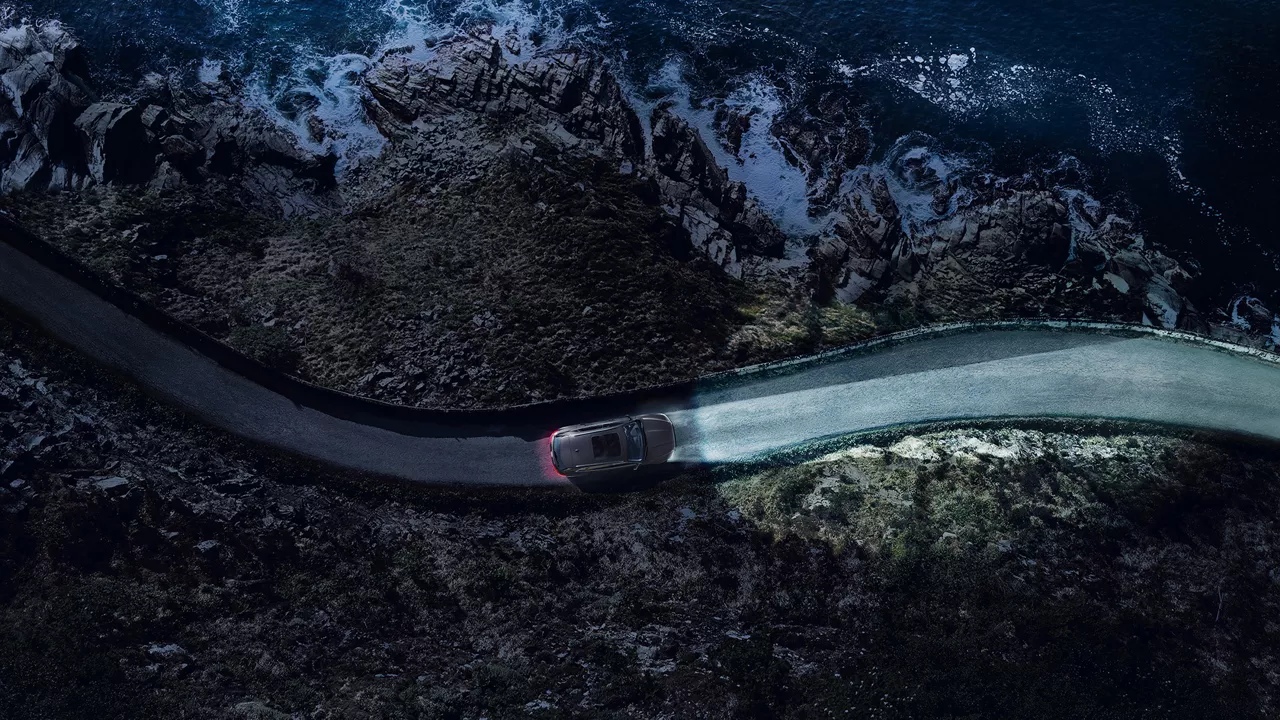 Auto mit eingeschaltetem Licht fährt bei Dunkelheit auf einer Küstenstraße entlang steiler Felsen und Wellen.