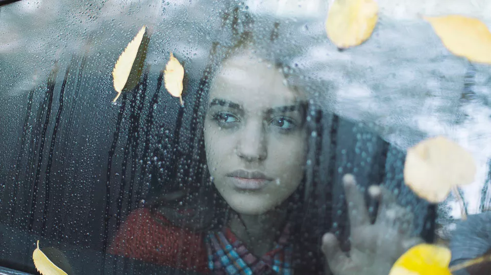 A woman stares out of a car window, with leaves and rain droplets decorating the surface of the vehicle.