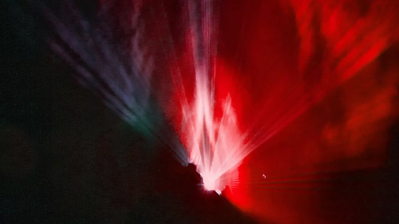 Ein Fotoausschnitt mit bunter Scheinwerferbeleuchtung. Auf der Linken Seite ist ein kräftiger roter Lichtstrahl zu sehen. Der nach recht über lila blau grün in die schwarze Dunkelheit verläuft. 