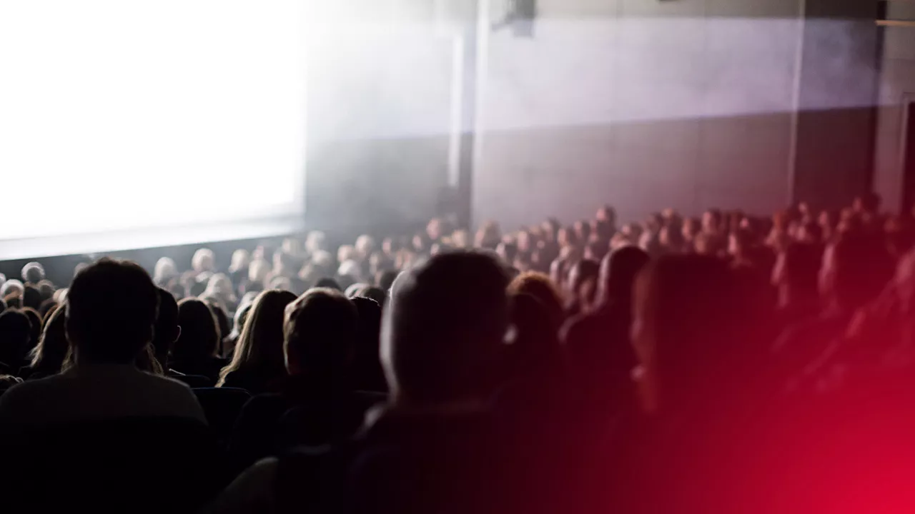 Ein großer Saal mit einer Leinwand von der ein heller weisser Lichtstrahl ausgeht. Das Publikum, das zur Leinwand blickt, ist von hinten zu sehen und nur schemenhaft aufgrund der diffusen Lichtverhältnisse zu erkennen.