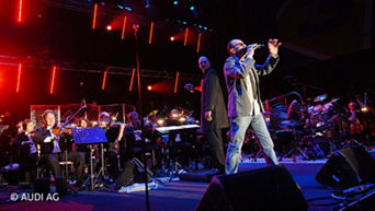Mann steht auf einer Bühne inmitten eines Liveorchestras und singt