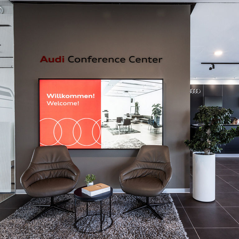 Empfangsbereich des Audi Conference Center mit zwei Stühlen und kleinem Tisch, im Hintergrund ein Display mit Willkommen 