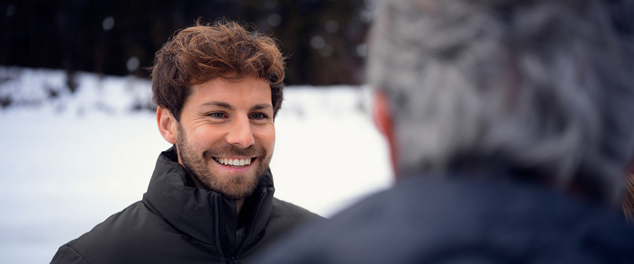 Mann mit braunen Haaren lächelt einen Mann mit grauen Haaren an (Grauhaariger Mann in Rückansicht), Hintergrund ist Schnee