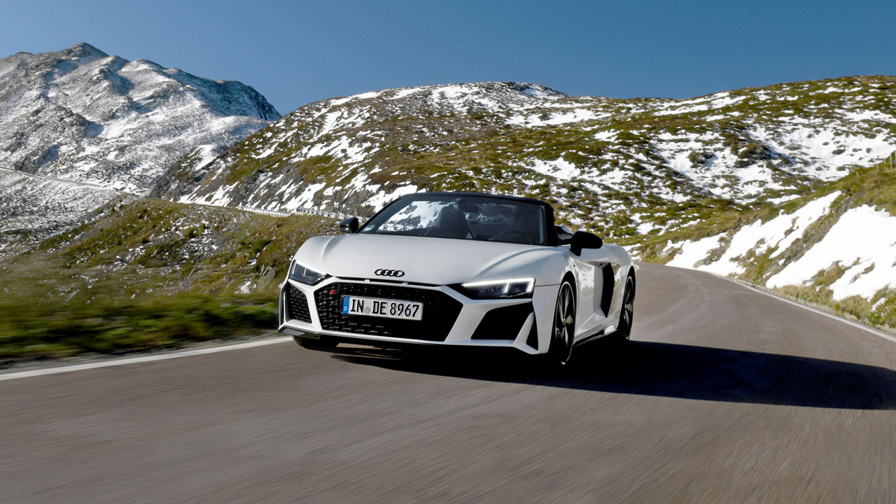 Weißer Audi R8 Spyder V10 performance quattro fährt auf einer Straße in den Bergen, die Berge sind mit Schnee bedeckt und der Himmel ist blau