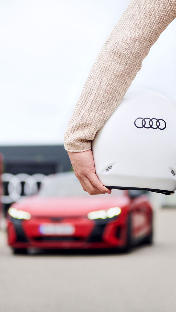 Rückansicht eines Mannes, der einen Helm mit Audi-Logo unter dem Arm trägt und auf ein rotes Audi Modell zugeht