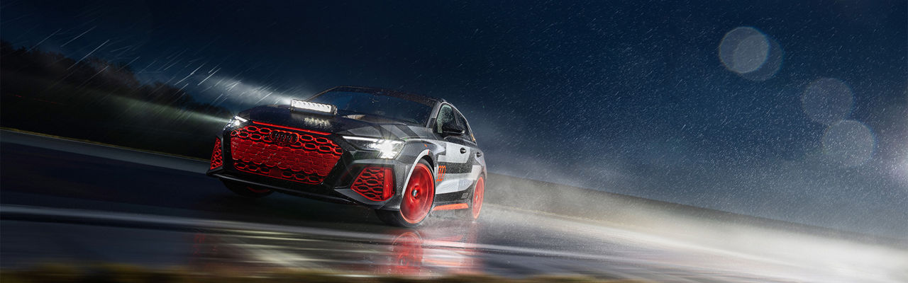 Audi RS 3 driving experience fährt auf nasser, dunkler Fahrbahn im Regen und hat die Lichter an