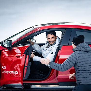 Ein Mann sitzt in einem roten Audi Modell auf der Fahrerseite mit geöffneter Tür. Ein weiterer Mann mit Mütze unterhält sich mit ihm