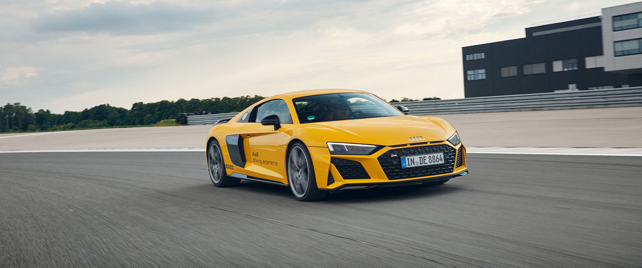 Gelber R8 Coupé V10 performance quattro mit schwarzer Aufschrift der Audi driving experience und Pirelli fährt auf Rennstrecke (Front- und Seitenansicht)