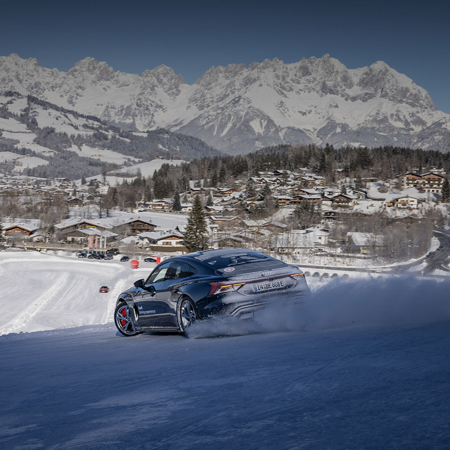 Dunkelgrauer Audi RS e-tron GT performance fährt auf einer Erhöhung im Schnee, im Hintergrund befindet sich weiter unten ein Parcour mit Audi Modellen, sowe Häusern und weiter hinten Berge 