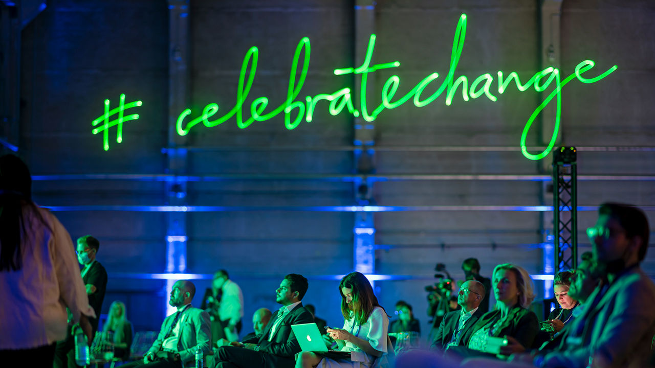 Le hashtag vert "#celebratechange" brille au-dessus des têtes des membres du public. 