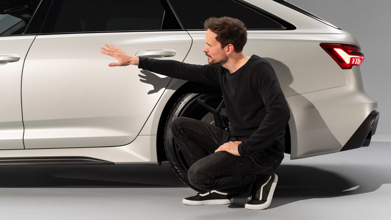 Markus Eberle accroupi à côté de l'Audi RS 6 Avant performance, fait un geste de la main en direction de la porte arrière.