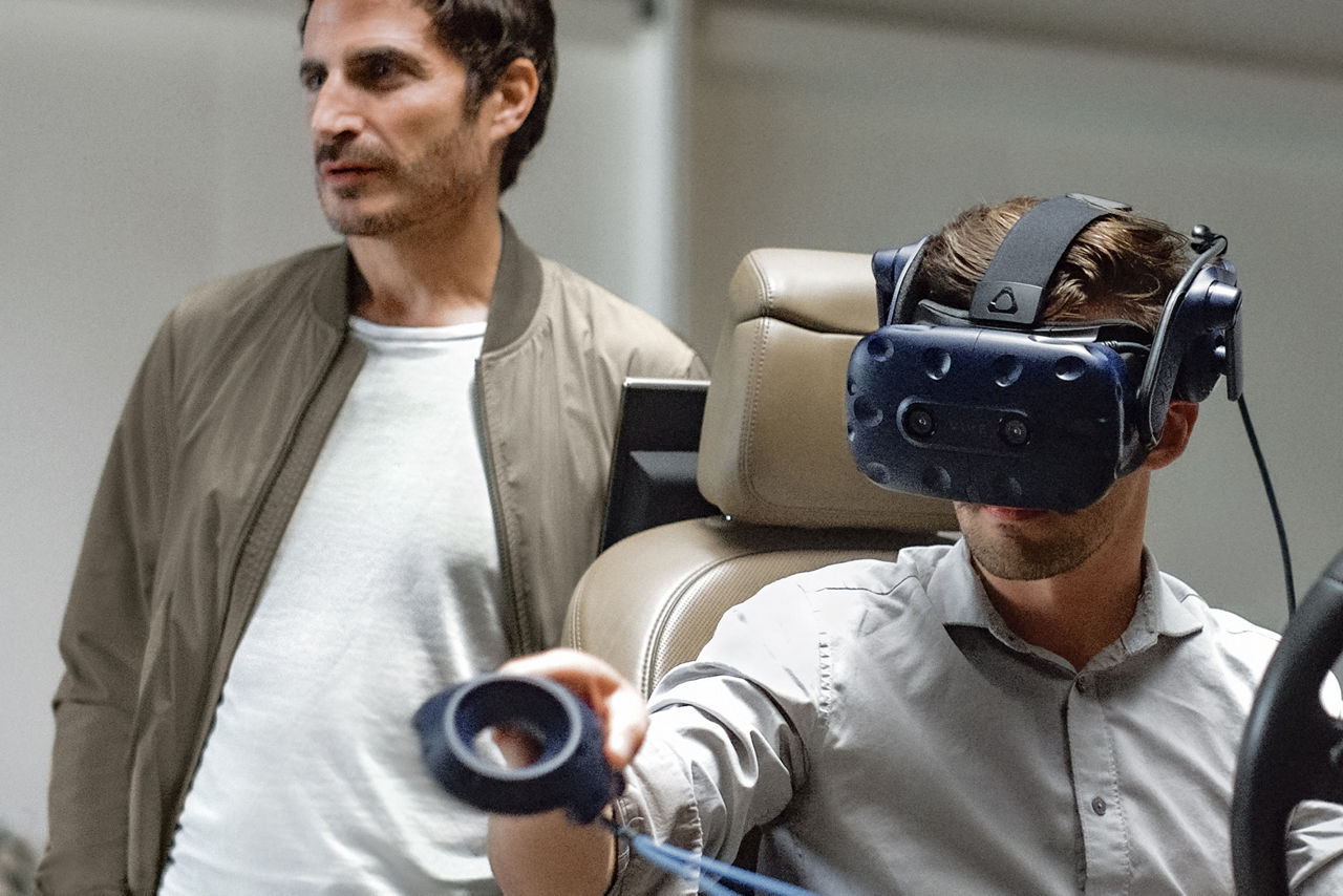 Gael Buzyn se tient derrière une personne assise dans un siège de voiture et portant des lunettes de réalité virtuelle.