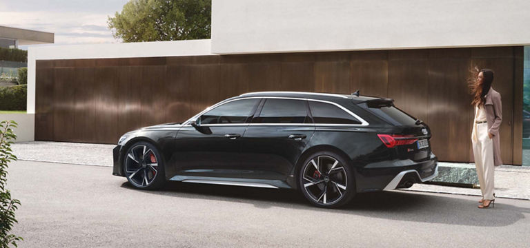 En Audi RS 6 Avant i Panther Black Crystal Effect foran et hus med en kvinne som står ved siden av bilen.