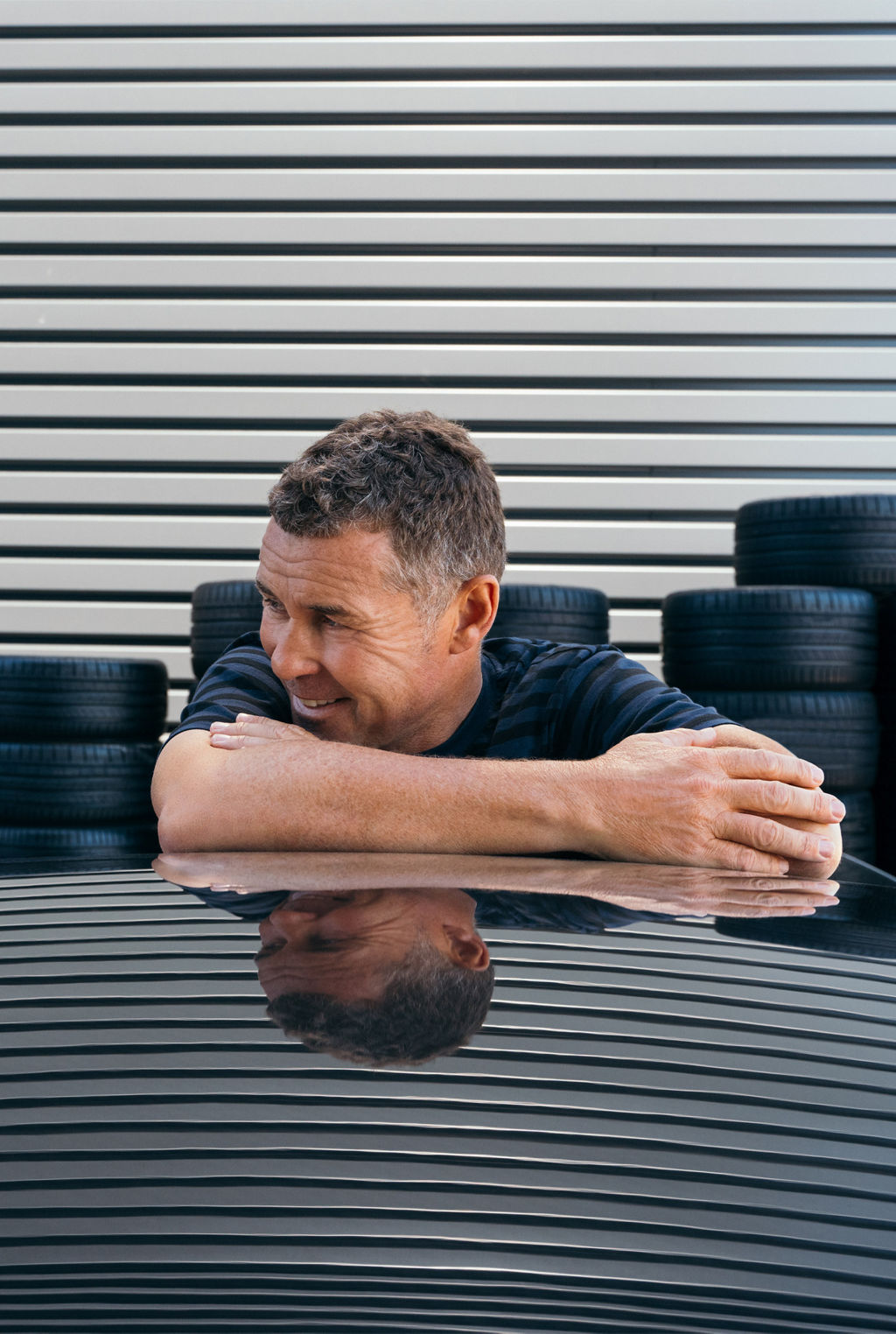 Tom Kristensen lehnt entspannt am Auto und lächelt verschmitzt zur Seite.
