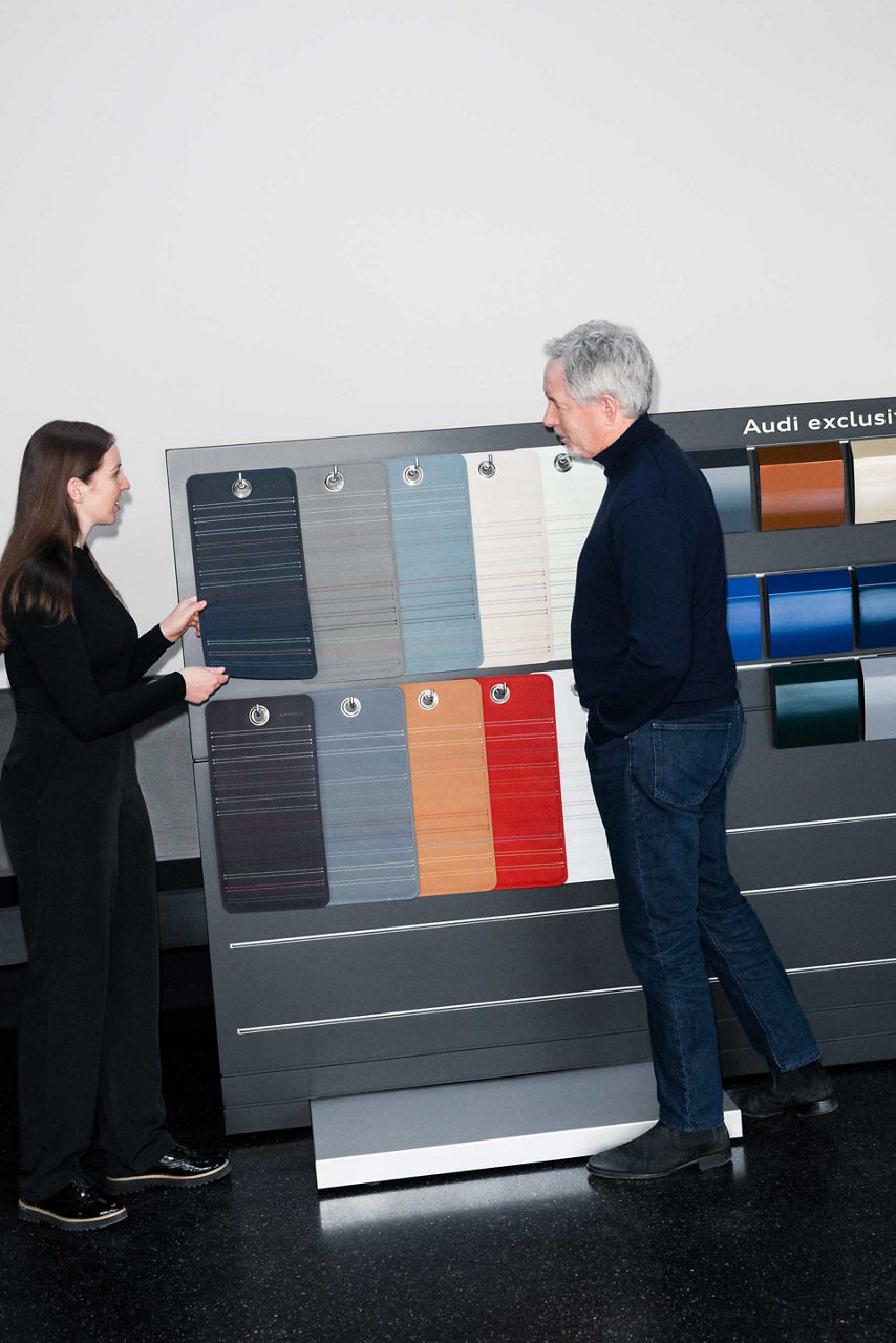 Annika Stößl und Trevor Hill unterhalten sich an der Audi exclusive Musterwand,  an der Interieur und Exterieur Farbmuster zu sehen sind. 
