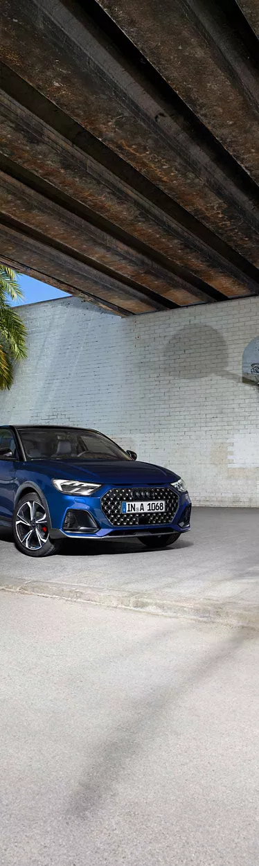 Blauer Audi A1 allstreet schräg von vorne links vor heller Ziegelwand mit sichtbarer Palmenpflanze.
