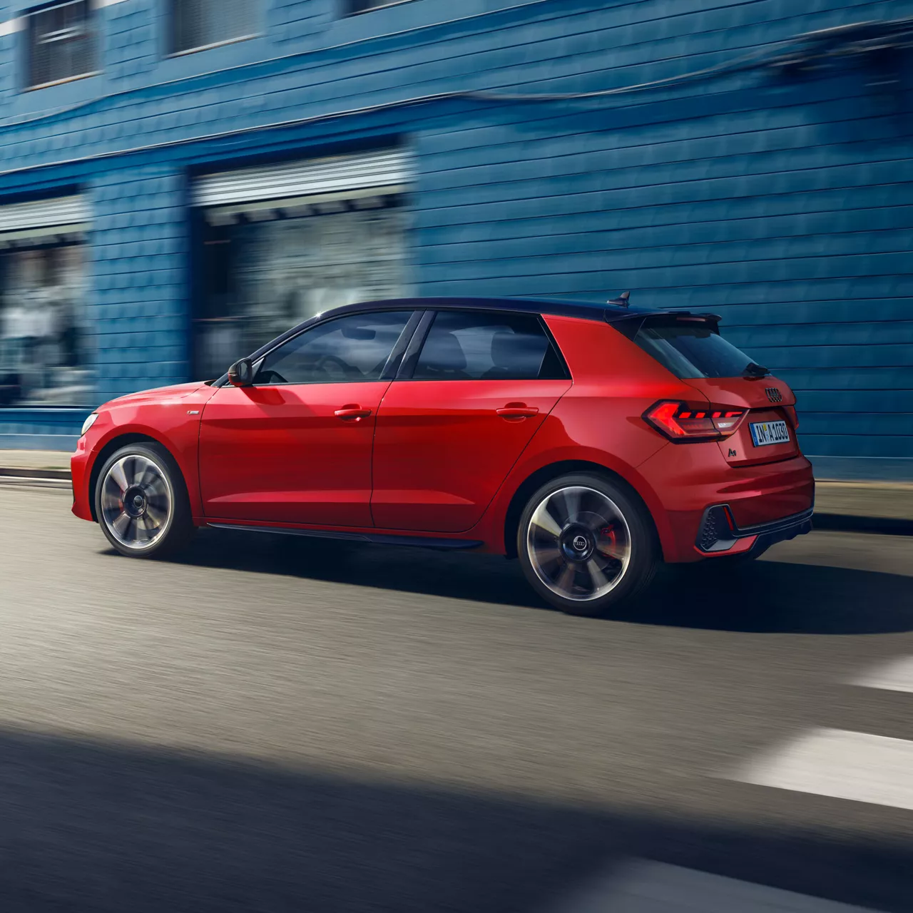 Roter Audi A1 Sportback fährt auf einer Straße vor einer blauen Wand, Seitenansicht von links.