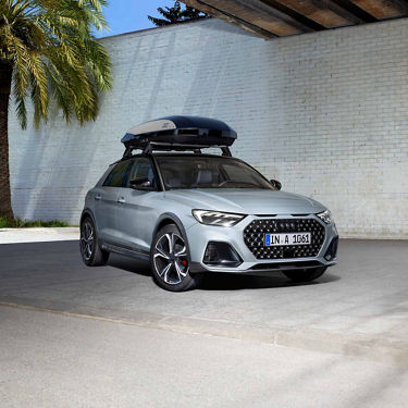 Silberner Audi A1 Sportback mit Dachbox, geparkt unter Brücke neben Palme und weißer Wand.