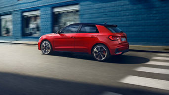 Ein roter Audi A1 Sportback fährt auf einer Straße mit Zebrastreifen, vorbei an einer blauen Gebäudefassade bei Tageslicht.