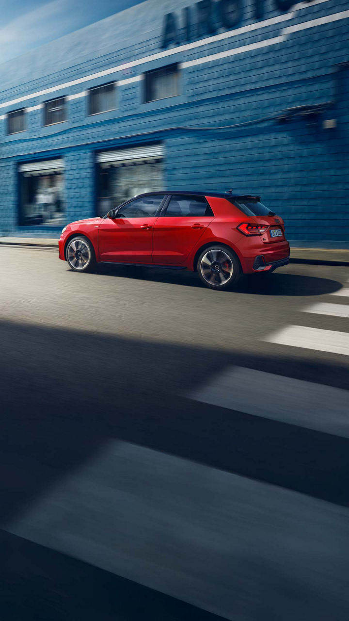 Roter Audi A1 Sportback fährt auf einer Straße vor einer blauen Wand, Seitenansicht von links.