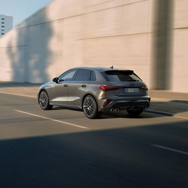 Rückansicht eines fahrenden, grauen Audi S3 Sportback auf leerer Straße bei Tageslicht.