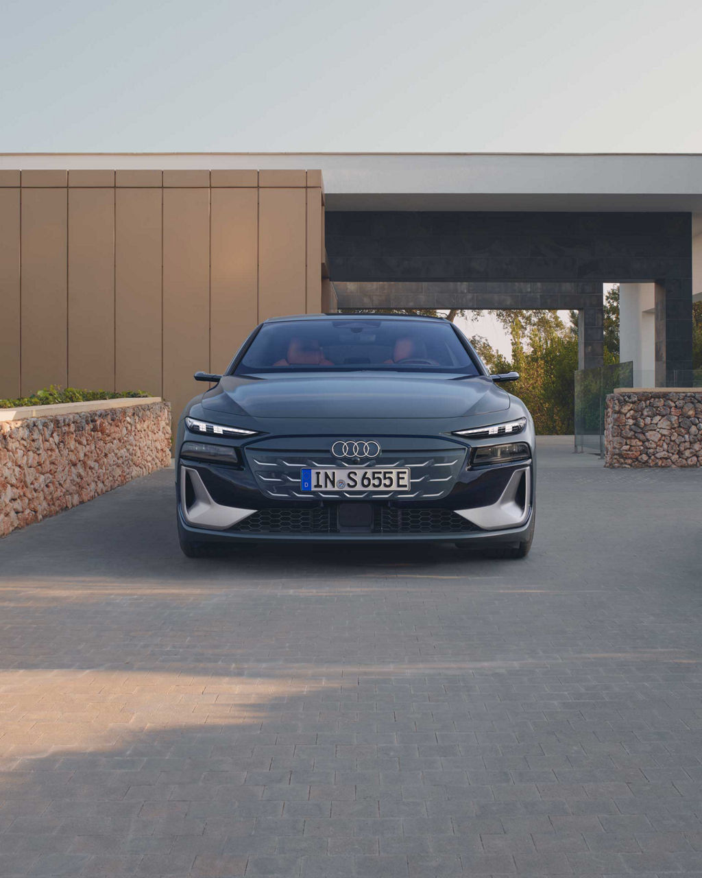 Ein dunkelblauer Audi S6 Avant e-tron steht frontal auf einer gepflasterten Einfahrt vor einem modernen Haus mit Natursteinmauer.