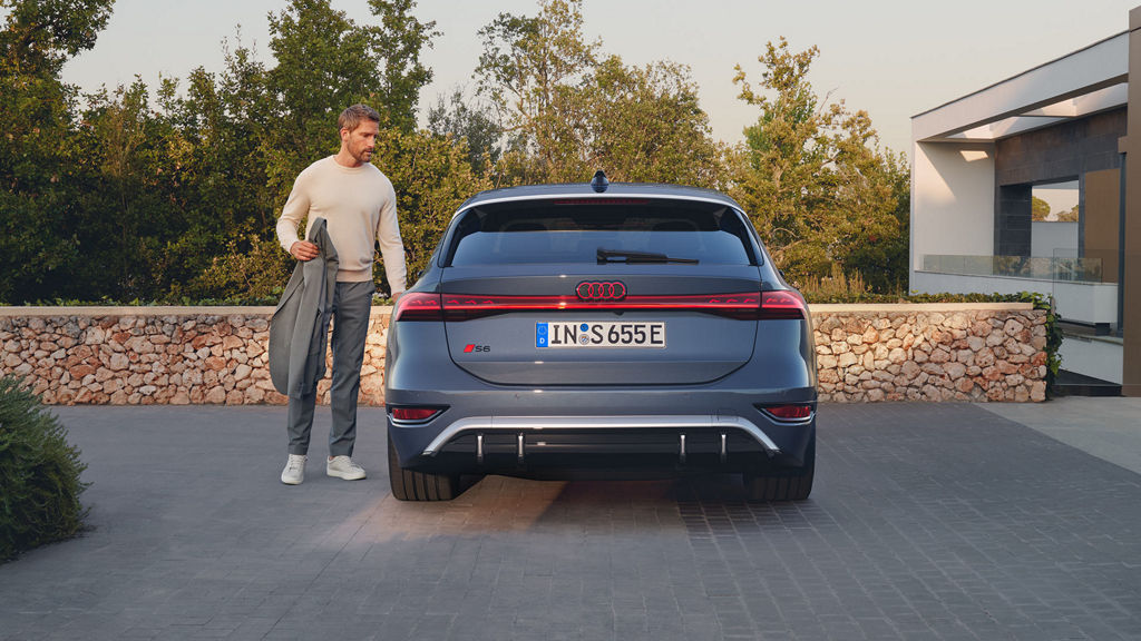 Ein Mann steht links neben einem dunkelblauen Audi S6 Avant e-tron, Rückansicht, auf einer gepflasterten Einfahrt vor einer Steinmauer.