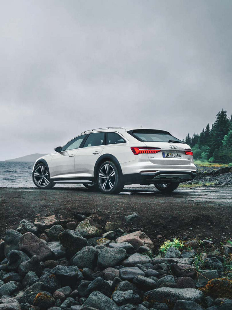 Weißer Audi A6 allroad quattro am Seeufer bei bewölktem Himmel, Blick auf Heck und Fahrerseite, auf nassem Untergrund geparkt.