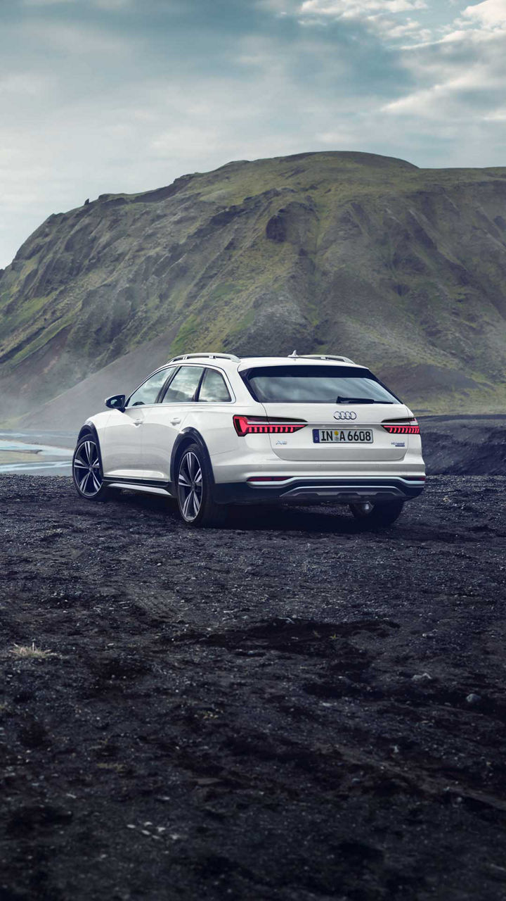 Weißer Audi A6 allroad quattro steht seitlich vor Bergkulisse auf Grasfläche neben einem ruhigen Gewässer, Ansicht schräg von hinten.