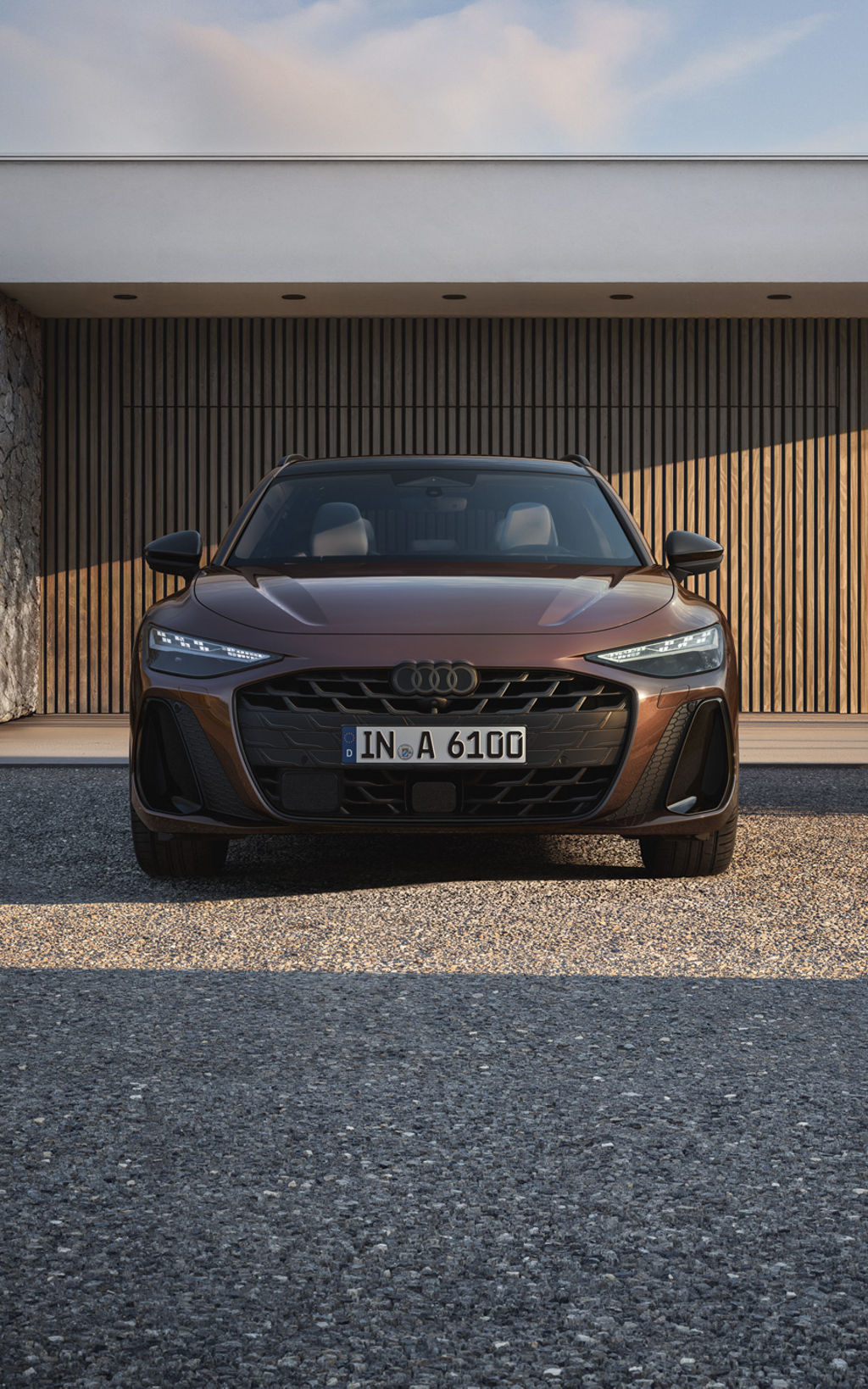 Brauner Audi A6 Avant Frontansicht, geparkt vor moderner Architektur mit Holz- und Steinfassade bei sonnigem Wetter.