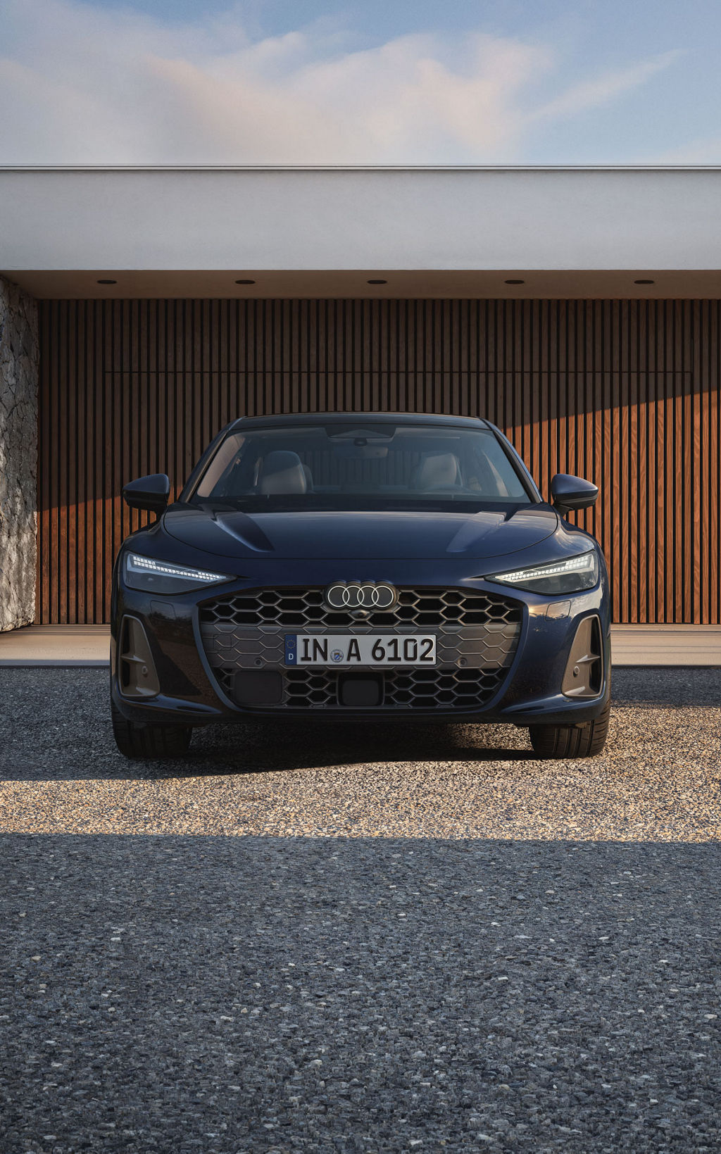 Die Frontansicht einer blauen Audi A6 Limousine, stehend auf einem Kiesuntergrund vor einem modernen Haus mit Holzverkleidung.