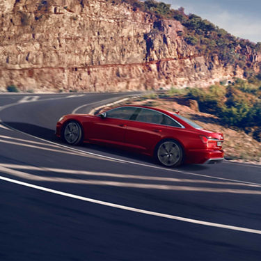 Rote Audi S6 Limousine TDI fährt dynamisch eine kurvige Bergstraße entlang bei sonnigem Wetter.