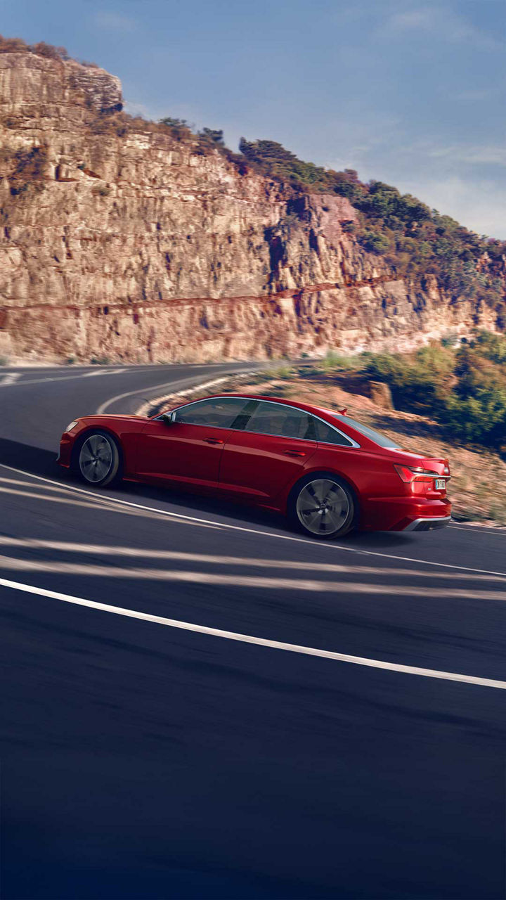 Rote Audi S6 Limousine TDI fährt dynamisch eine kurvige Bergstraße entlang bei sonnigem Wetter.