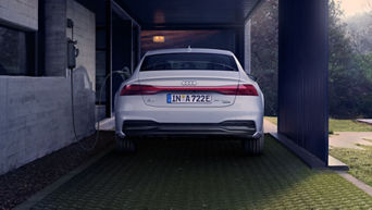 Heckansicht eines weißen Audi A7 Sportback TFSI e, der unter einem Carport per Kabel an eine Wallbox angeschlossen ist.