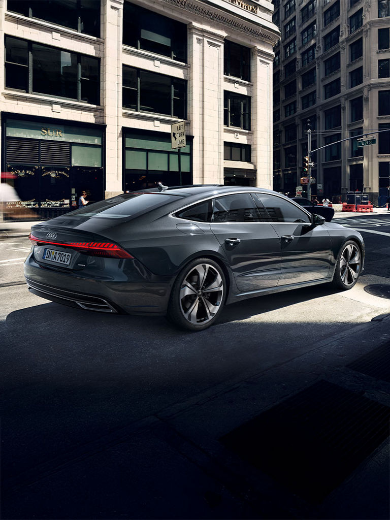 Dunkelgrauer Audi A7 Sportback fährt durch eine Stadtstraße, umgeben von Gebäuden und Verkehr, seitliche Rückansicht.