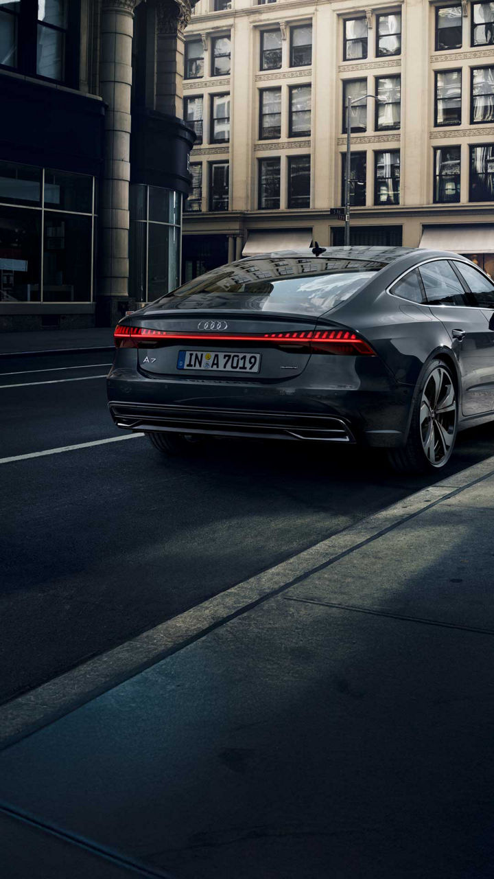 Dunkelgrauer Audi A7 Sportback hält auf städtischer Straße, Fußgänger überqueren Zebrastreifen im Hintergrund.
