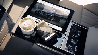 Ein Kaffee, ein Tablet und ein Handy liegen auf einer Mittelkonsole im Auto