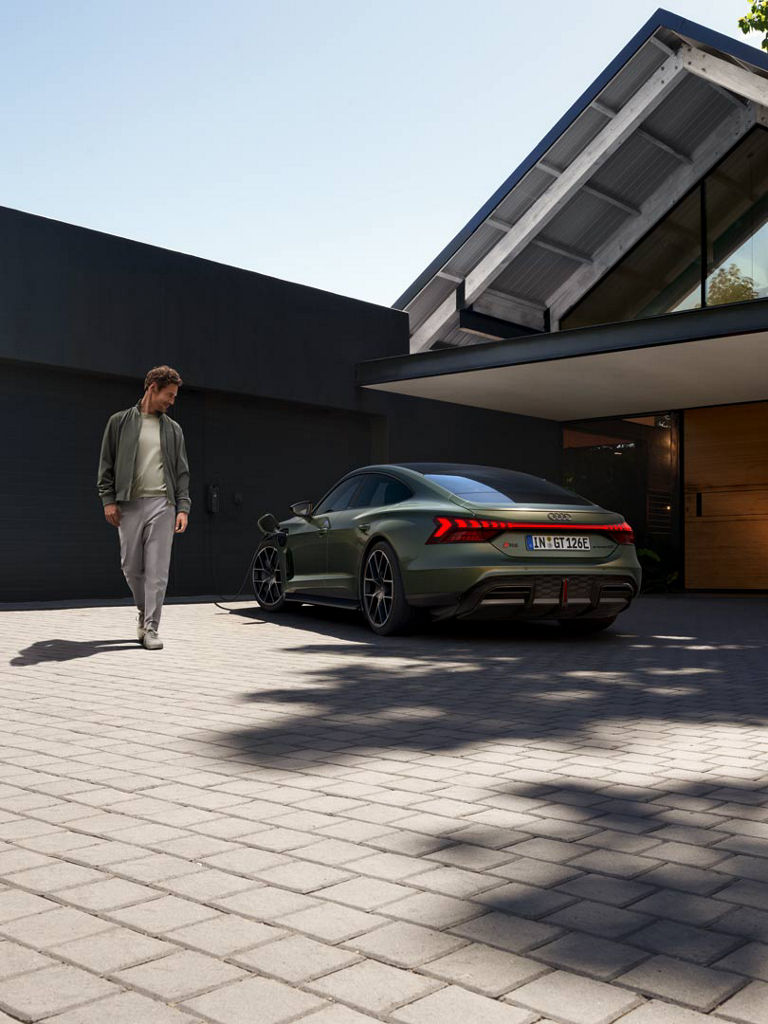 Grüner Audi RS e-tron GT performance lädt an einer Wallbox vor einem modernen Haus, ein Mann geht am Fahrzeug vorbei.