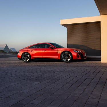 Ein roter Audi S e-tron GT ist an einer modernen Ladestation neben einem Haus angeschlossen, mit einer minimalistischen Architektur und einer ruhigen Landschaft im Hintergrund.